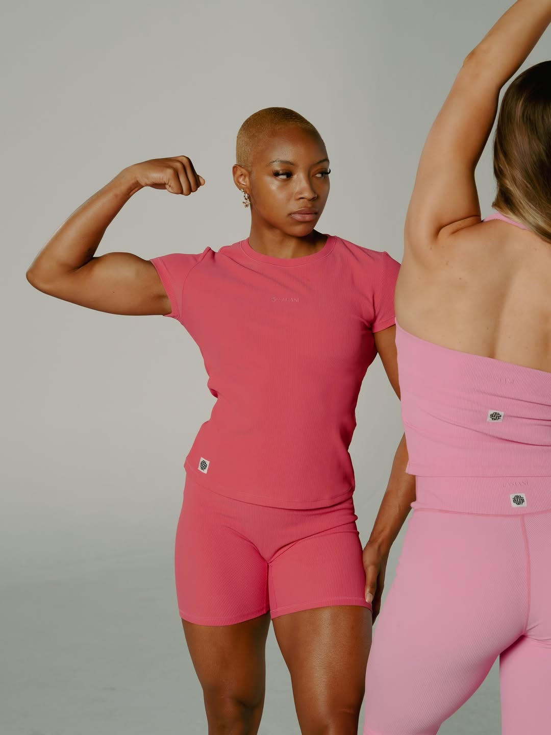 Three-quarter fitness shot flexing a bicep in a pink ribbed set, second model behind
