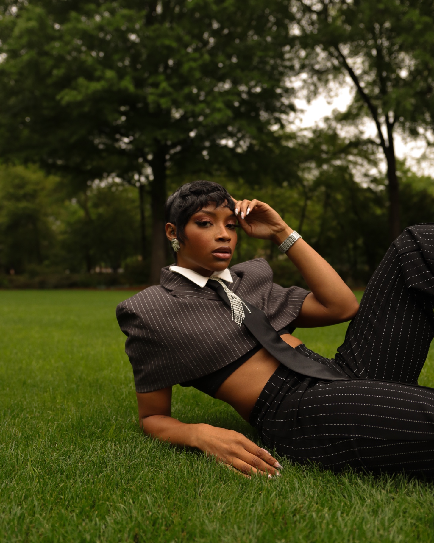 Reclining editorial fashion shot in a cropped pinstripe suit with a rhinestone tie on a lawn