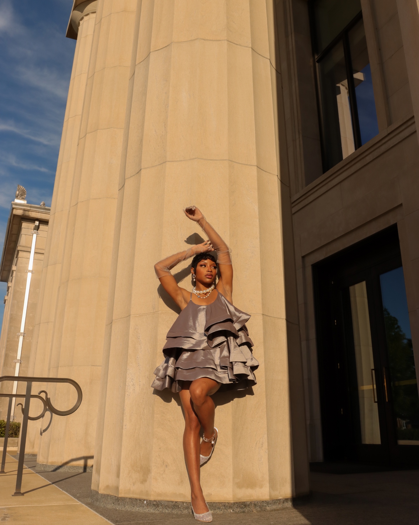 Full-length fashion editorial in a metallic ruffle dress, arms raised, against stone columns