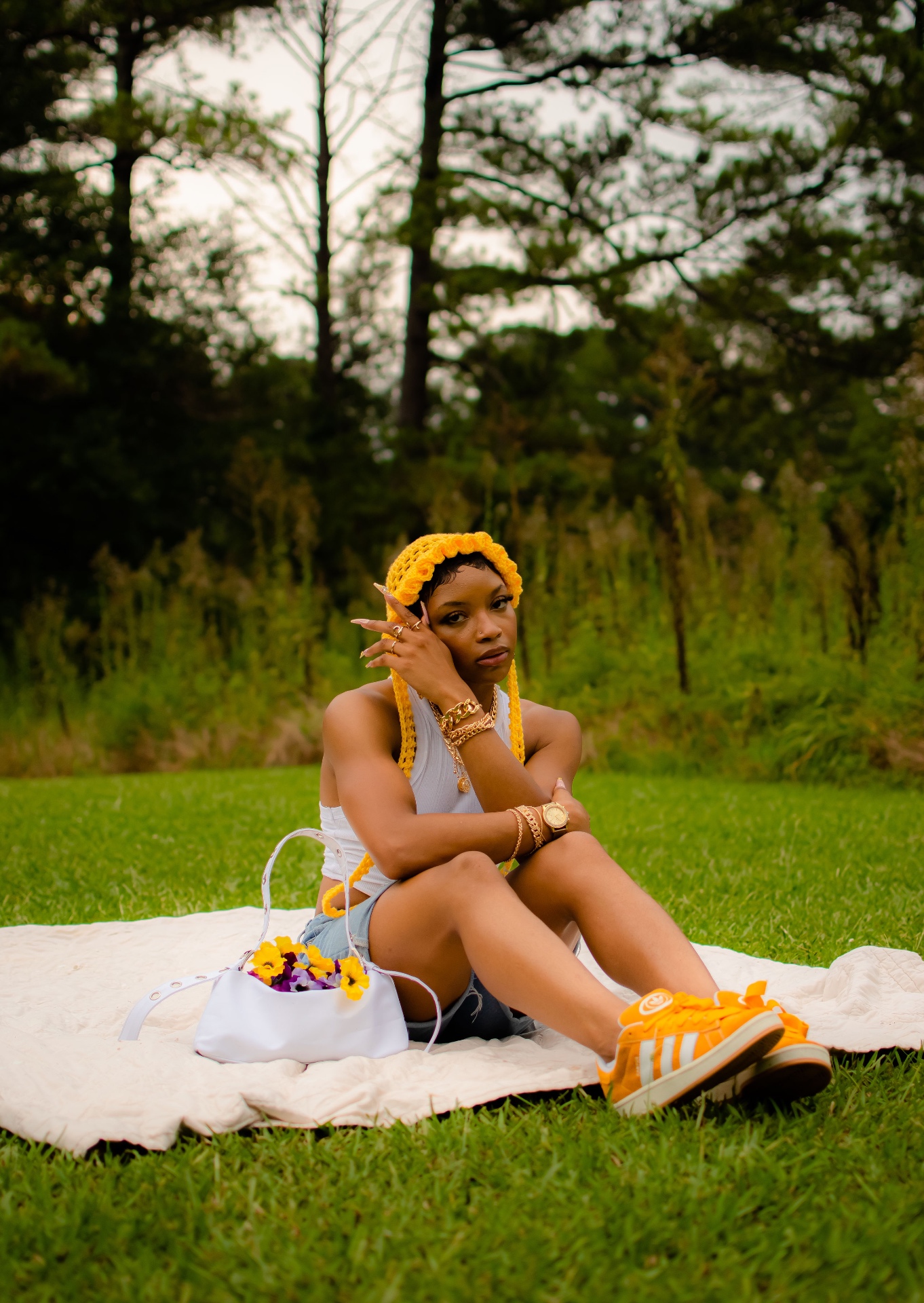 Editorial fashion shot in a yellow knitted headpiece, white tank and denim shorts sitting on a blanket in tall grass