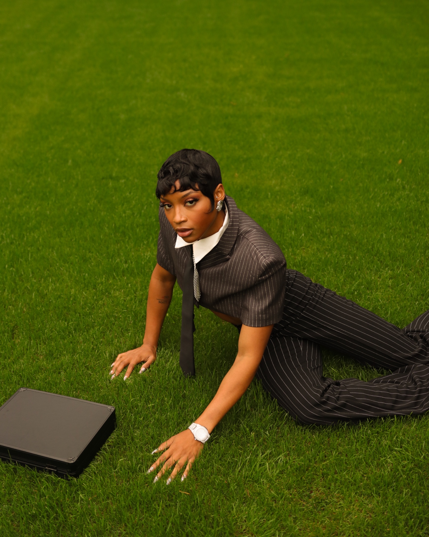 High-angle editorial fashion shot in a pinstripe suit on a lawn beside a briefcase