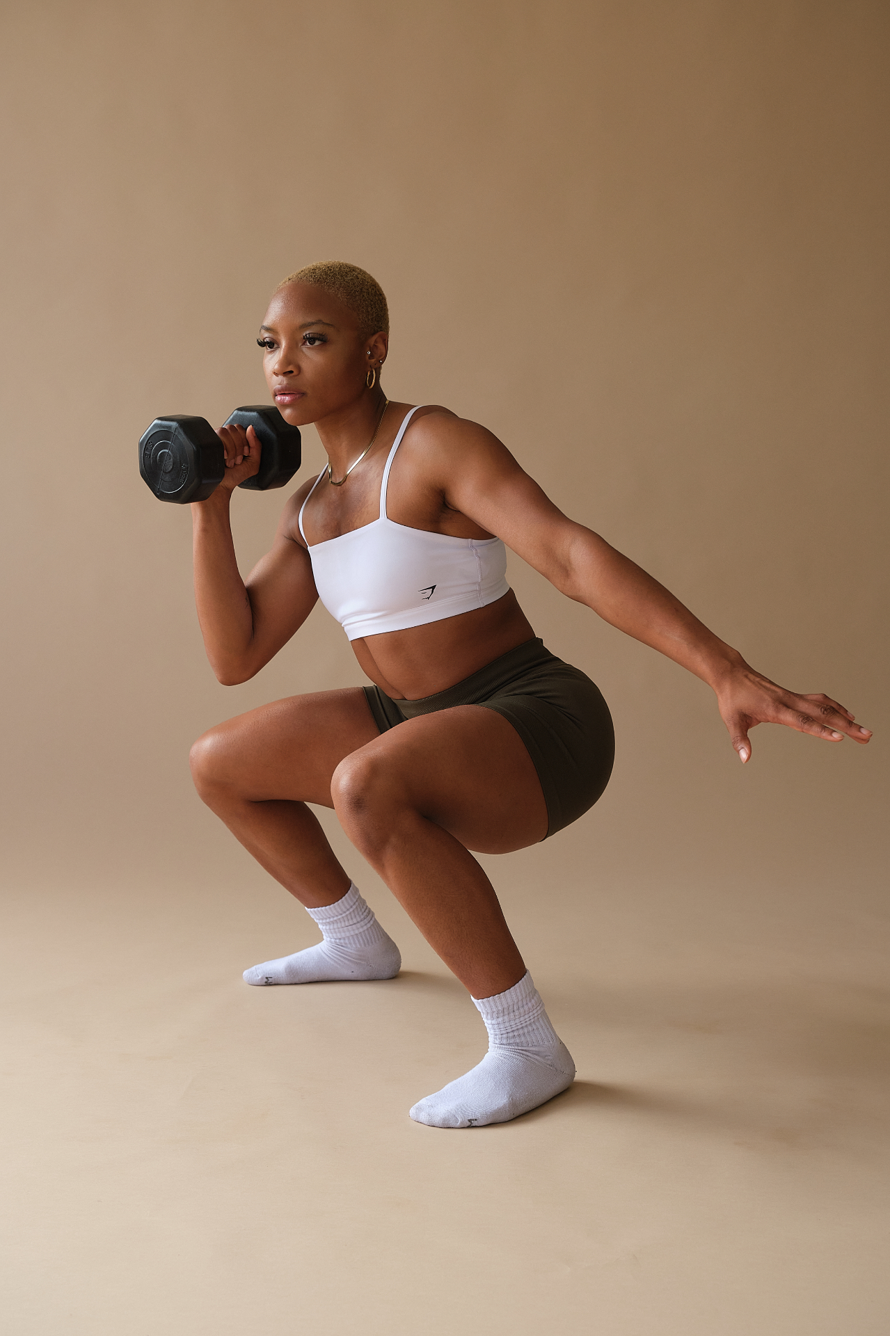 Full-length fitness shot squatting with a dumbbell against a warm tan studio backdrop