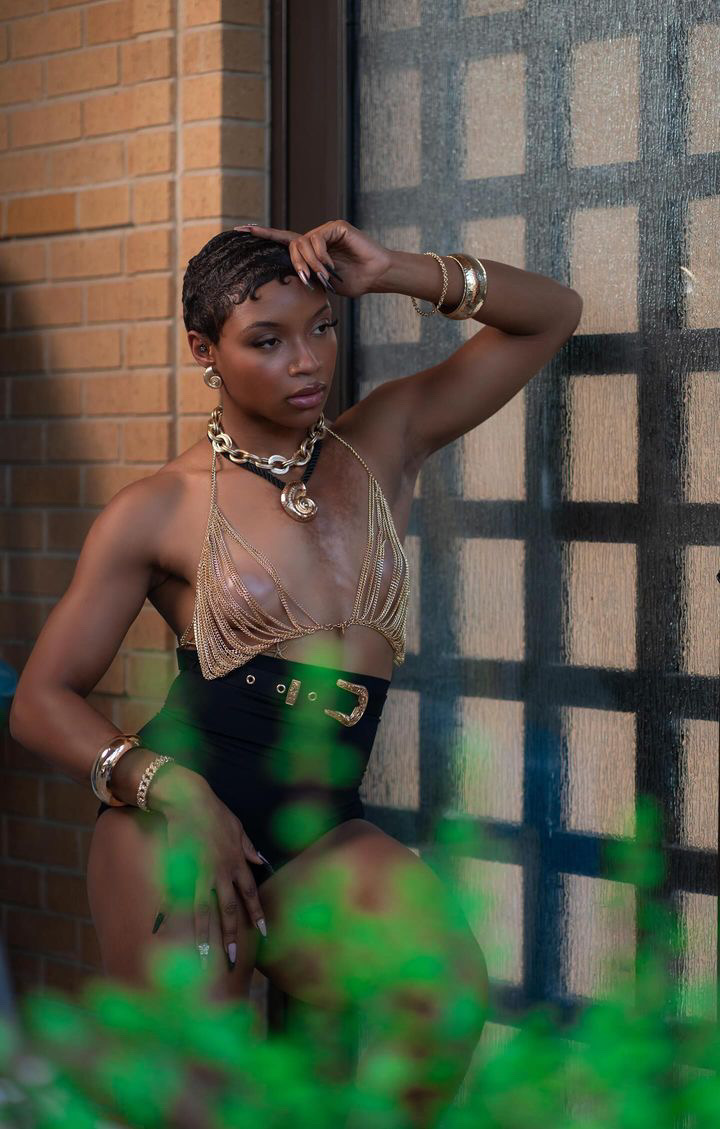 Editorial fashion shot in a gold chain halter top and black shorts against a brick wall