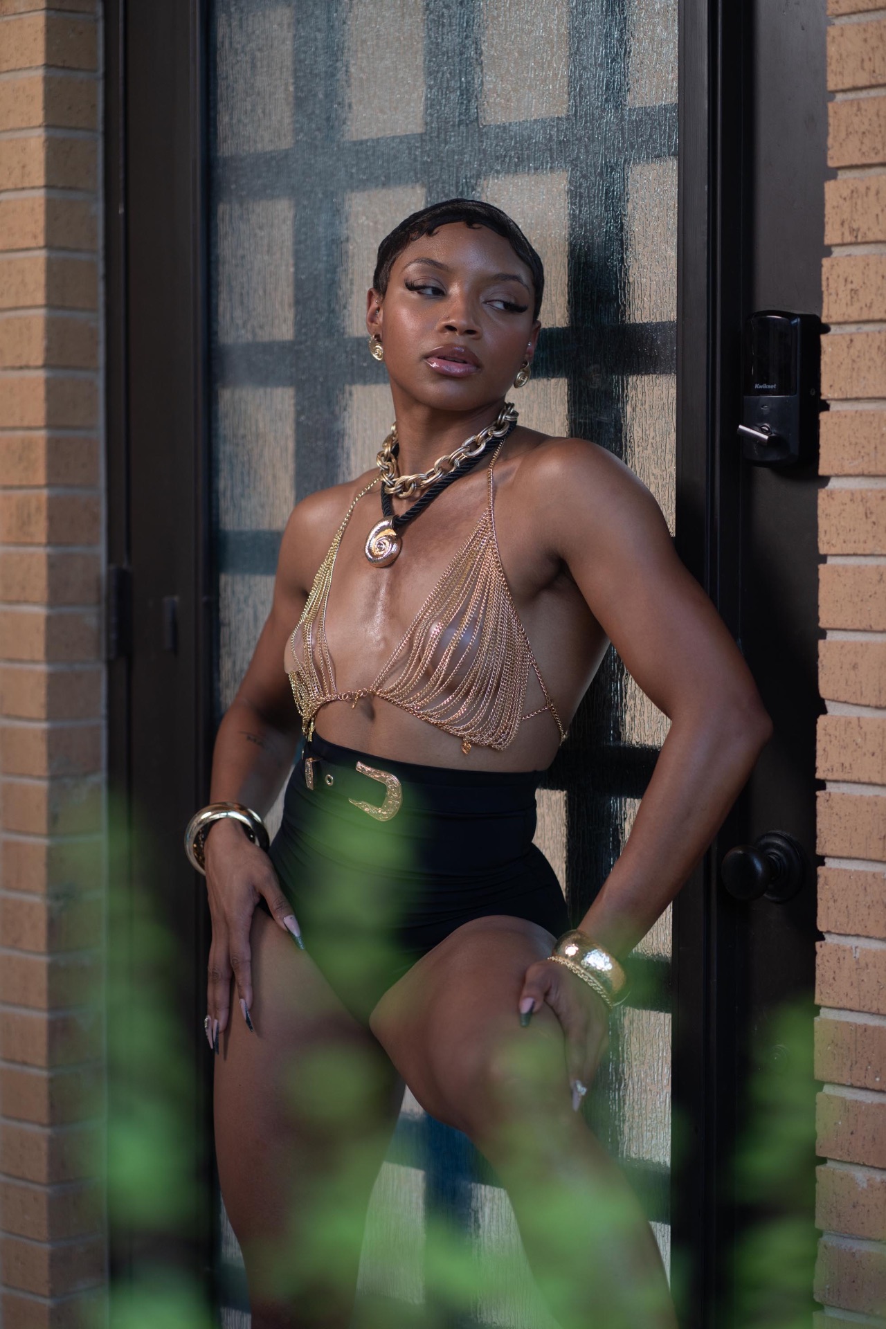 Editorial fashion shot in a gold chain halter top and black high-waisted shorts leaning on a glass door
