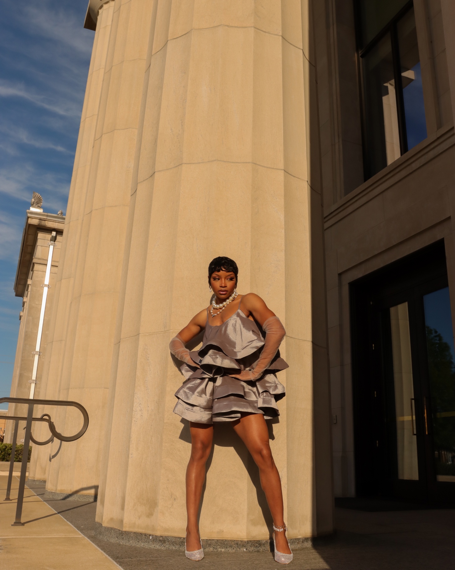 Full-length fashion editorial in a metallic tiered ruffle dress and opera gloves against stone columns